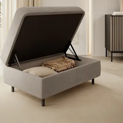 Nowoczesna pufa Gomsi Touch 18 z funkcją schowka – podnoszone wieko na czarnych siłownikach ułatwia odkładanie koców i poduszek. Pufa stoi na dywanie w jasnym salonie, obok komody Meorati i rośliny w donicy przy zasłonach.