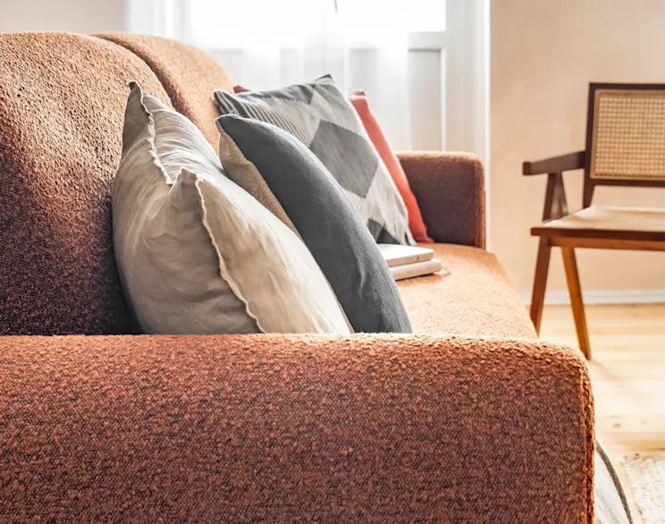 Zbliżenie na kanapę Arcadova w tkaninie Jaffray 30, ozdobioną poduszkami w różnych kolorach, w jasnym i przytulnym wnętrzu z krzesłem z rattanu.