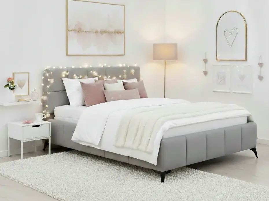 Łóżko Mist Nube 03 jasnoszare 180x200 z tapicerowanym zagłówkiem w nowoczesnej sypialni. Na łóżku znajduje się biała kołdra i poduszki w odcieniach pudrowego różu, a całość dopełnia miękki jasny dywan.