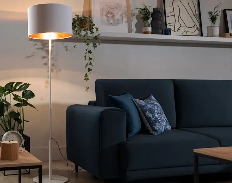 Lampa stojąca Vespillo z białym abażurem i złotym wnętrzem świeci ciepłym światłem obok granatowej sofy. W tle widać półkę z dekoracjami, rośliny doniczkowe oraz drewniany stolik kawowy, co tworzy przytulną, nowoczesną aranżację salonu.