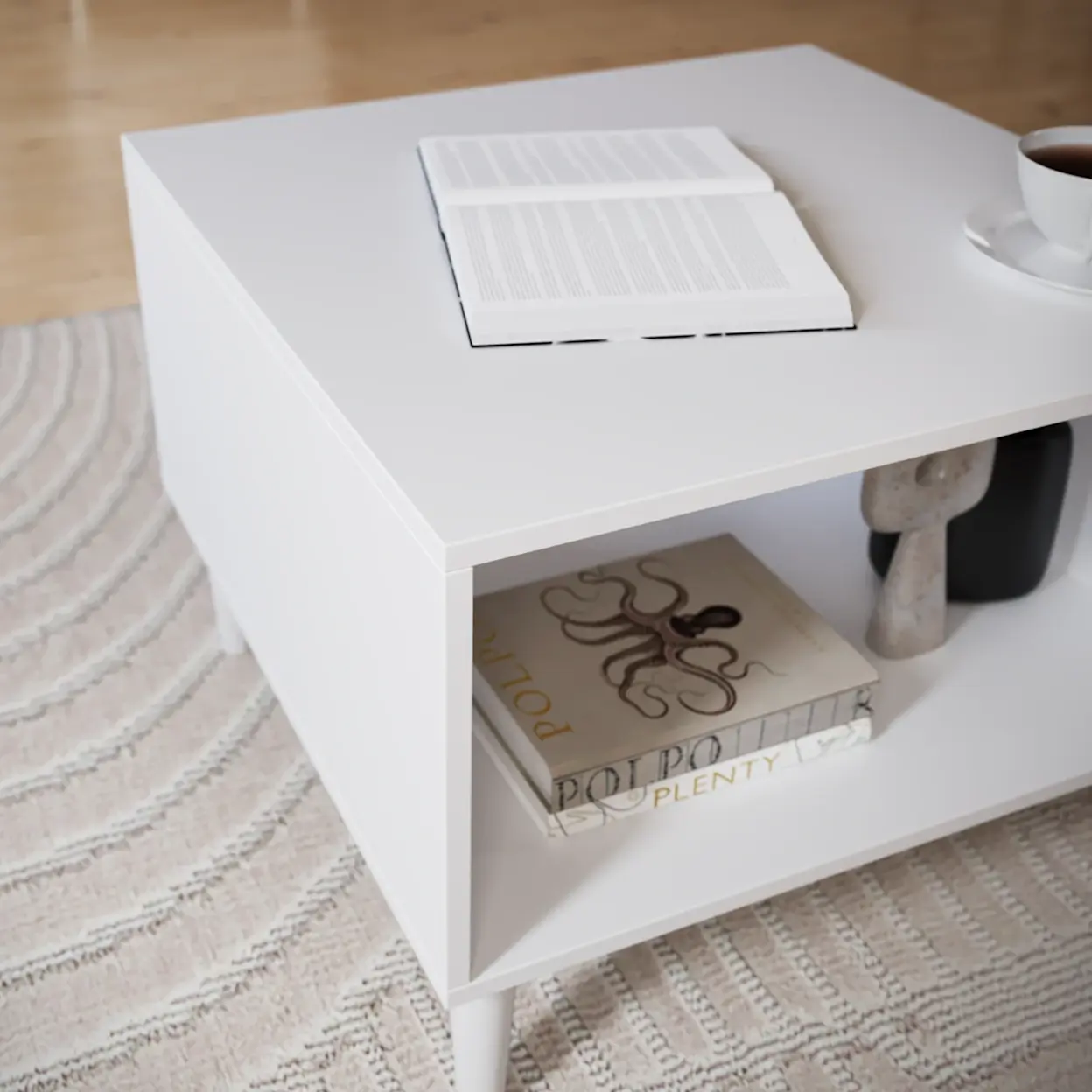 Biały stolik kawowy Dullum o prostej, minimalistycznej formie z praktyczną półką pod blatem. Na górze leży otwarta książka i filiżanka kawy, a na dolnej półce widać albumy i dekorację.