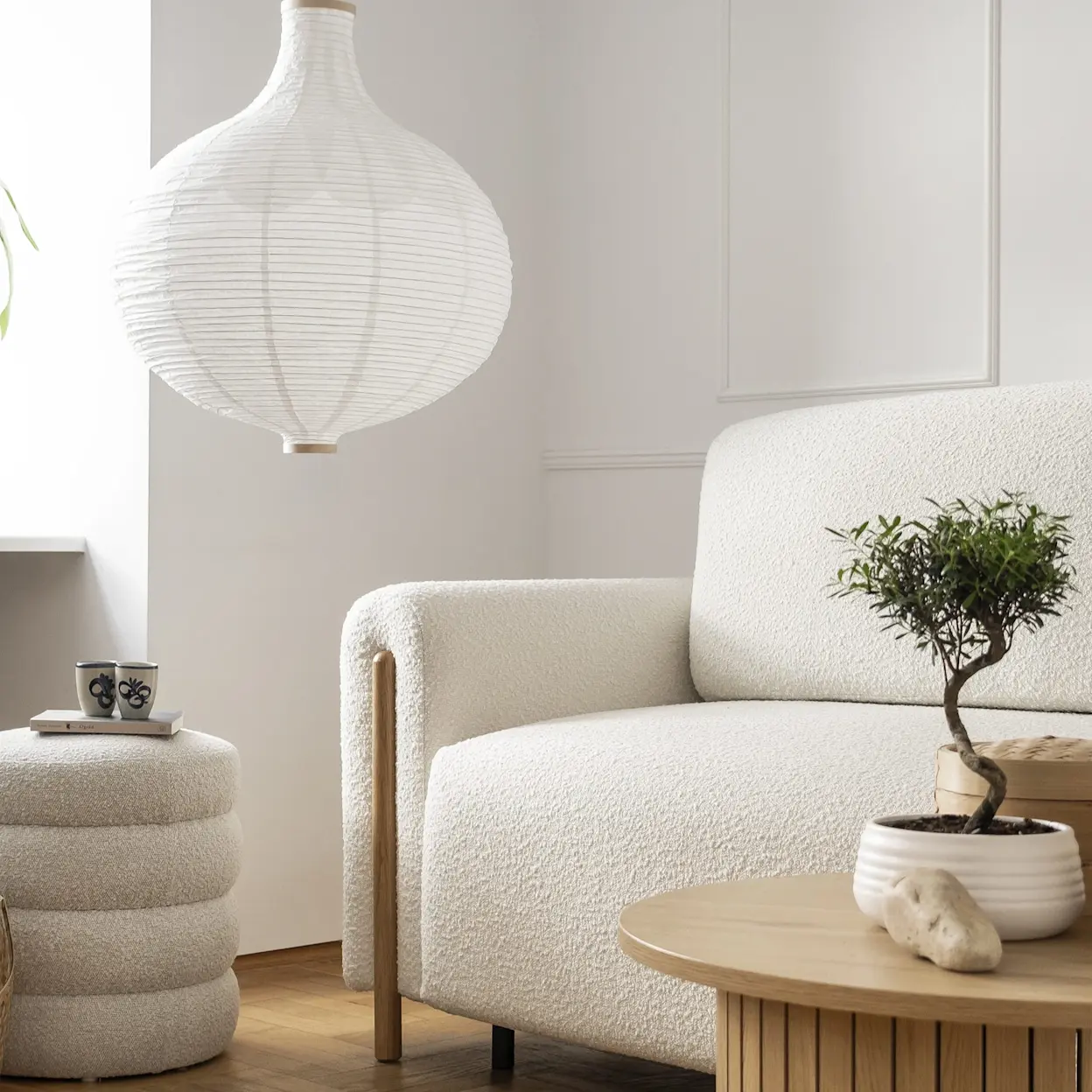 Jasna kanapa Arcadova w tkaninie boucle w minimalistycznym salonie w stylu japandi. Obok stoi okrągły stolik z drewnianym wykończeniem i rośliną, a wnętrze rozjaśnia papierowa lampa.