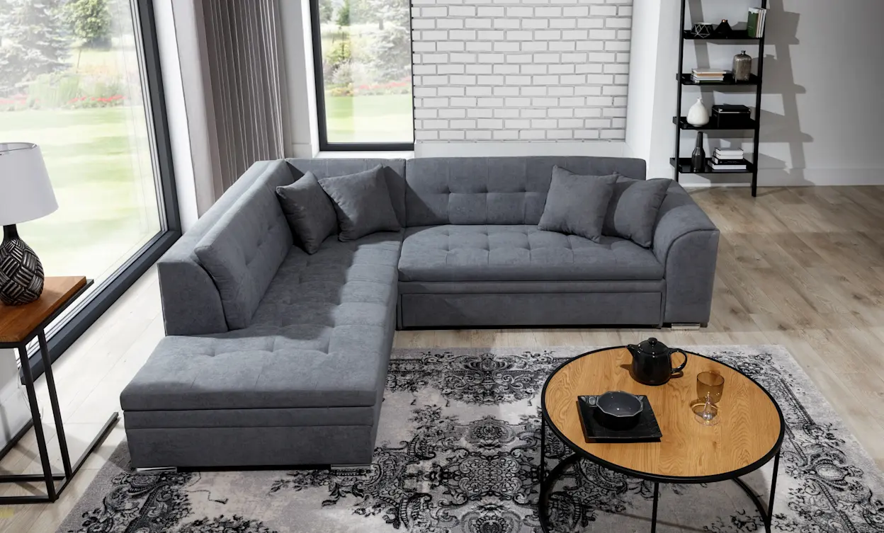 Szary narożnik Pieretta w przestronnym, jasnym salonie z dużymi oknami, ozdobiony poduszkami i ustawiony na dekoracyjnym dywanie.