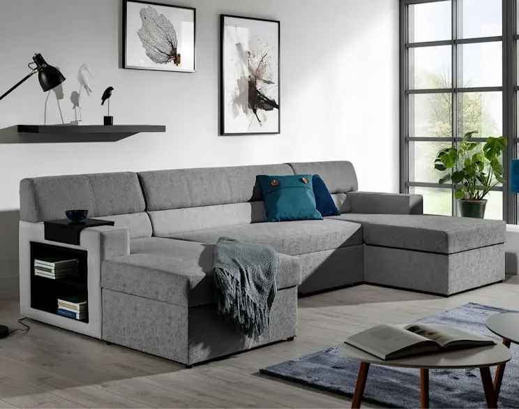 Szary narożnik Markos z boczną półką i dekoracyjnymi poduszkami, ustawiony w jasnym salonie z dużymi oknami i minimalistycznymi dekoracjami.