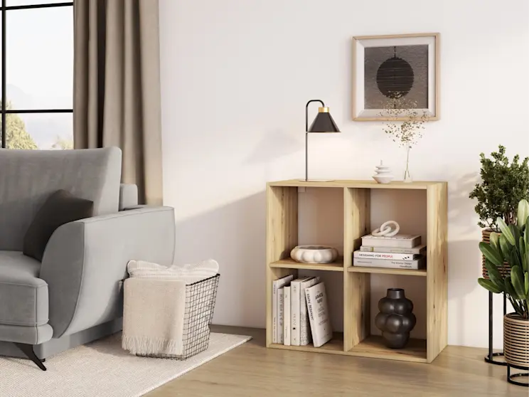 Drewniany regał Jugis z otwartymi półkami ustawiony przy ścianie, obok fragment kanapy Megis Monolith 84 w jasnym kolorze. Na regale widać lampkę Detme, książki i dodatki, które podkreślają nowoczesny, minimalistyczny styl wnętrza.