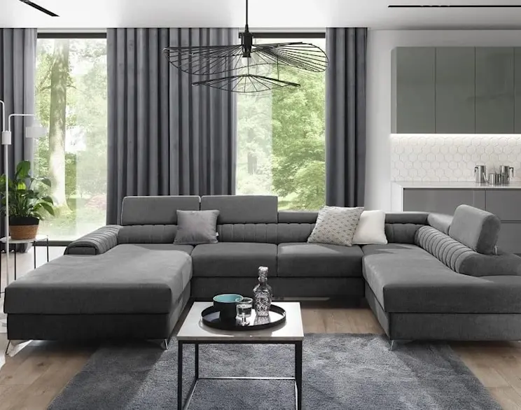 Nowoczesny salon utrzymany w szarej tonacji z szarym narożnikiem i widokiem na zieleń za dużymi oknami. Wnętrze charakteryzuje minimalistyczny design