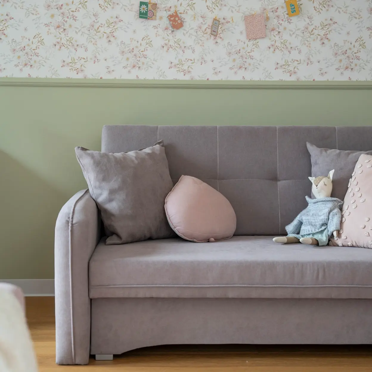 Szara kanapa dla dzieci Laine w pokoju dziecięcym – tapicerowana sofa z zaokrąglonym podłokietnikiem, poduszkami i pluszową zabawką. W tle zielona lamperia oraz delikatna, kwiatowa tapeta.