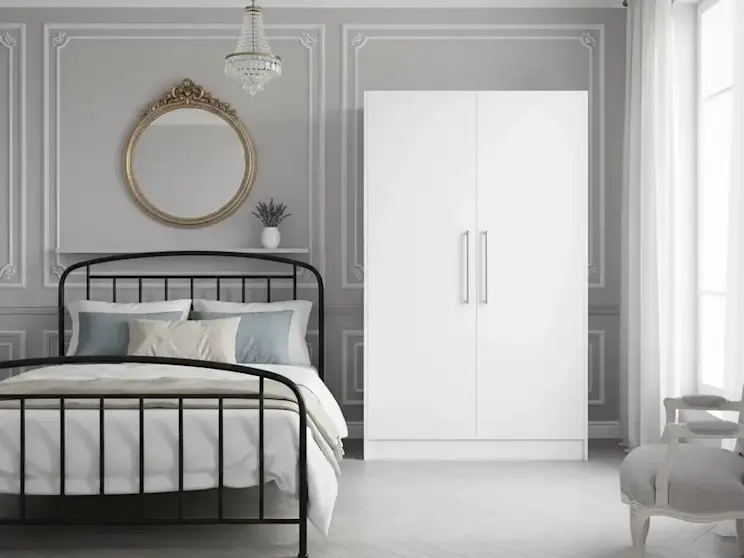 Szafa Alarica II w kolorze białym ustawiona w jasnej sypialni o klasycznym charakterze, z ozdobnym żyrandolem, tapicerowanym krzesłem i dużym oknem. Mebel idealnie wpisuje się w elegancką aranżację wnętrza.