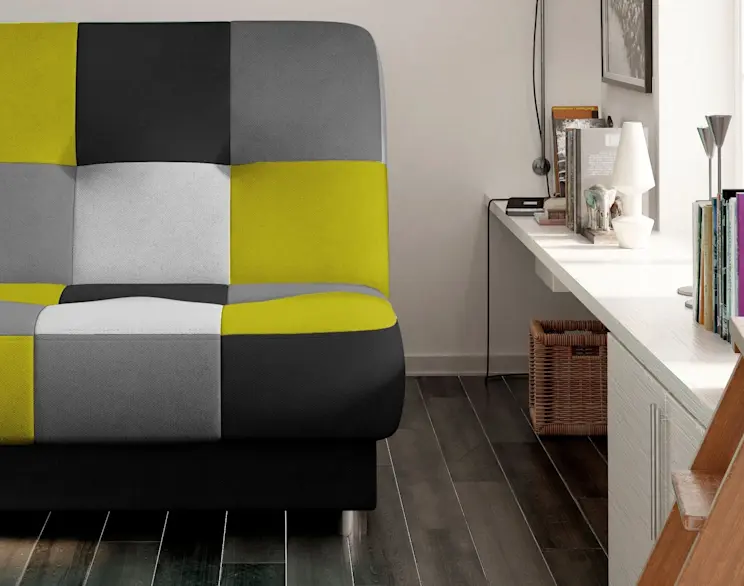 Wersalka Cayo w stylu patchwork: żółto-szaro-czarna tapicerka z kontrastowymi panelami. Mebel stoi w jasnym pokoju obok białego biurka z książkami i lampką, na ciemnej drewnianej podłodze.