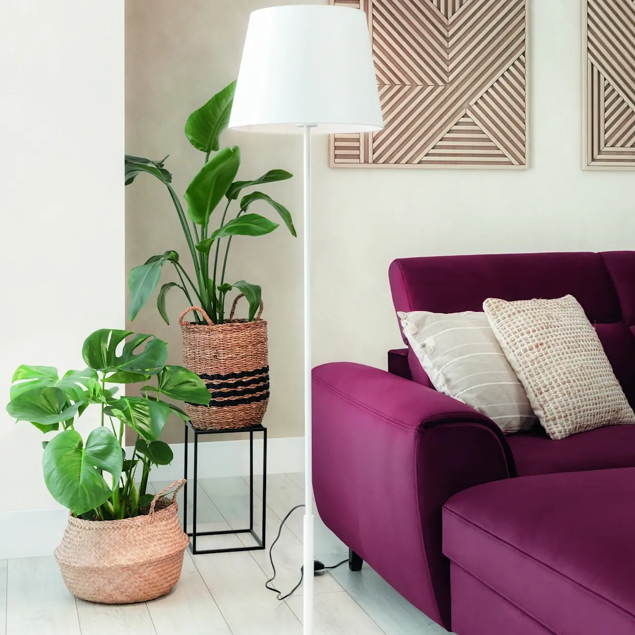 Biała lampa stojąca Vespillo z dużym, jasnym abażurem stoi w salonie obok fioletowej sofy. Przy lampie są zielone rośliny w plecionych koszach, a całość tworzy przytulny, domowy kącik.