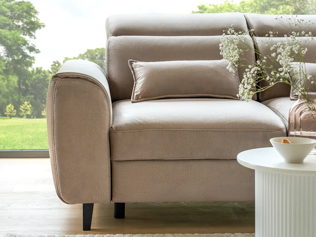 Detal narożnika Foble w jasnym beżu, z eleganckimi przeszyciami i dekoracyjną poduszką, ustawiony przy oknie z widokiem na ogród.