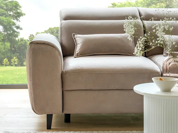 Detal narożnika Foble w jasnym beżu, z eleganckimi przeszyciami i dekoracyjną poduszką, ustawiony przy oknie z widokiem na ogród.