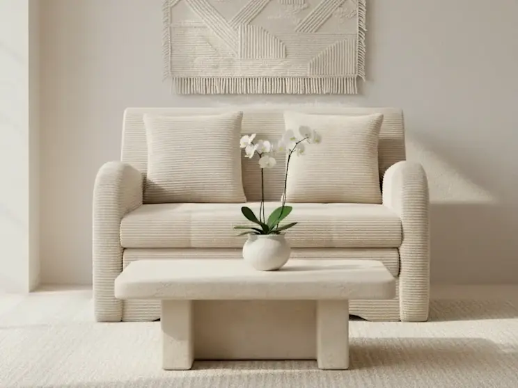 Beżowa sofa Ario w tkaninie Jarell 18 ustawiona w minimalistycznym salonie z jasnymi ścianami, stolikiem kawowym z kamiennym blatem oraz dekoracyjną makramą na ścianie, tworząca spokojną i harmonijną aranżację wnętrza.