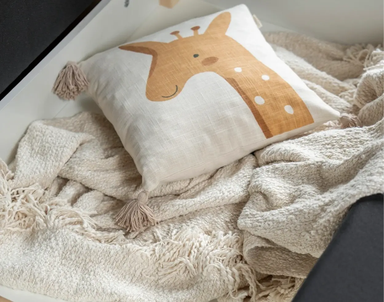 Zbliżenie na jasną poduszkę z żyrafą i chwostami ułożoną na miękkim, beżowym kocu. Kadr pokazuje przytulne tekstylia w aranżacji pokoju dziecka przy sofie Ario z funkcją spania i pojemnikiem na pościel.