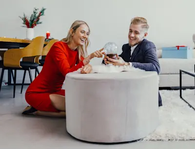 Para siedząca przy okrągłej pufie Bero w jasnym salonie, trzymająca kulę śnieżną, w tle dekoracje świąteczne i sofa w tkaninie boucle.