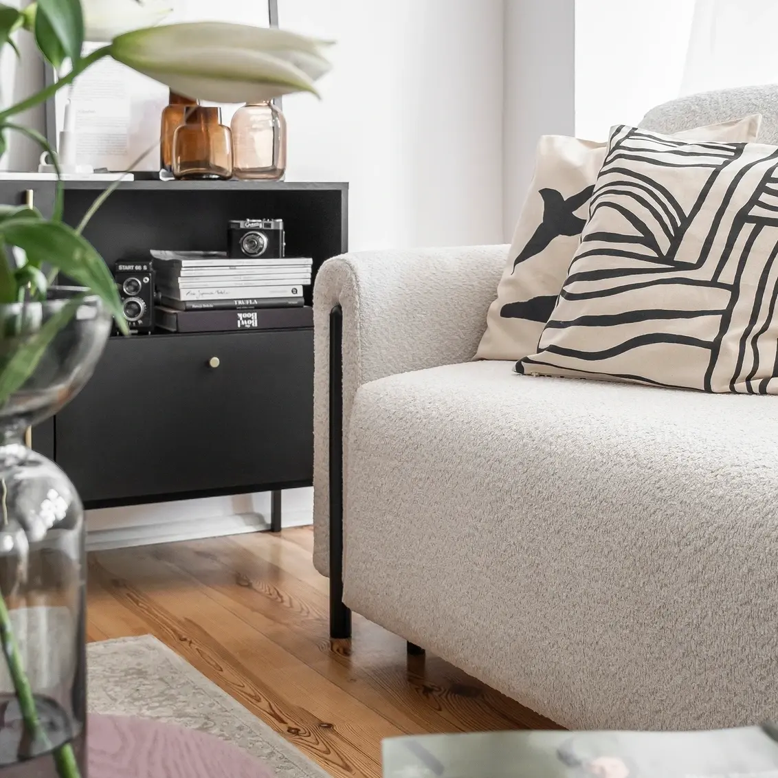 Jasna kanapa Arcadova II w przytulnym salonie z poduszkami w czarno-białe wzory. Obok stoi czarna komoda Lanzzi z półką, na której ułożono książki i dekoracje, a całość dopełnia jasne światło z okna.