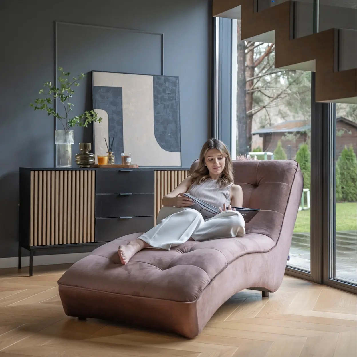 Aranżacja salonu z wygodnym szezlongiem Loren o pikowanym siedzisku i opływowej formie, na którym odpoczywa osoba z magazynem. Obok widoczna jest komoda Meorati z ryflowanym drewnem, dekoracjami i obrazem, podkreślając przytulny klimat.