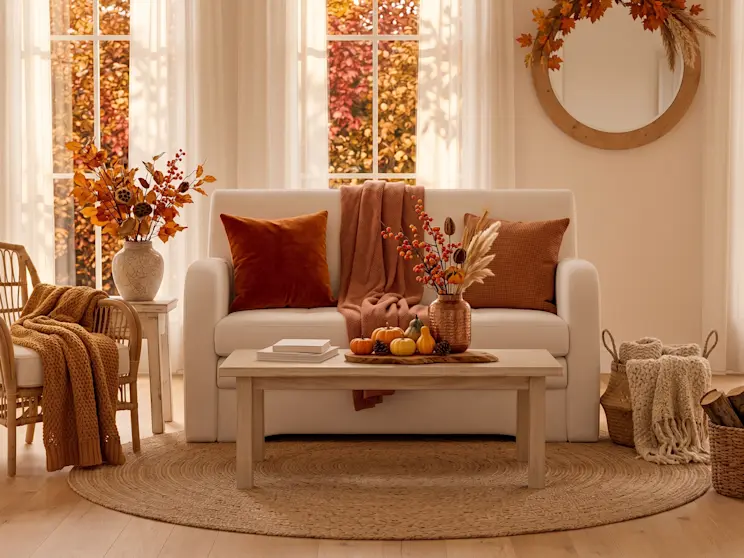 Biała sofa Ario Royal 01 prezentowana w eleganckim salonie z rattanowym fotelem, drewnianym stolikiem kawowym oraz dekoracyjnym lustrem ściennym. Jasna kolorystyka i naturalne materiały tworzą harmonijną aranżację.