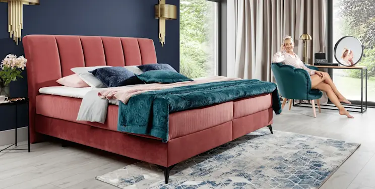 Tapicerowane łóżko Aderito w tkaninie Kronos 29 w różowym kolorze, z wysokim pionowo pikowanym wezgłowiem. Aranżacja sypialni z granatową ścianą, turkusowym pledem, jasną podłogą i dywanem we wzór.