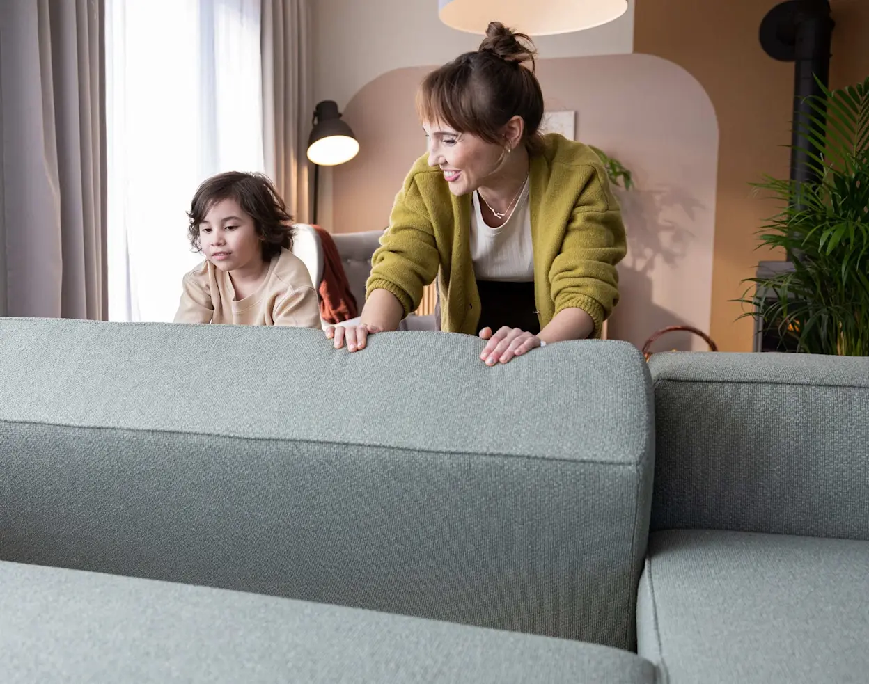 Wiosenne porządki w salonie z narożnikiem rozkładanym Aurumm – mama i dziecko poprawiają poduszki i oparcie na szarej sofie, w tle lampy i rośliny. Przytulna aranżacja dla rodzinnego wnętrza.