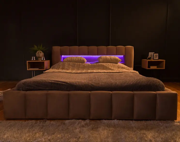 Fragment aranżacji sypialni: tapicerowane łóżko Lamica Monolith 09 z zaokrąglonym zagłówkiem i efektowną listwą LED świecącą na fioletowo. Po lewej widoczny stolik nocny z ozdobną donicą w kształcie głowy i świecznikami na tle czarnej ściany.