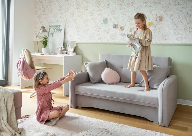 Szara amerykanka Laine w tkaninie Poco 03 w pokoju dziecięcym, z bawiącymi się dziewczynkami i dekoracjami w pastelowych kolorach.