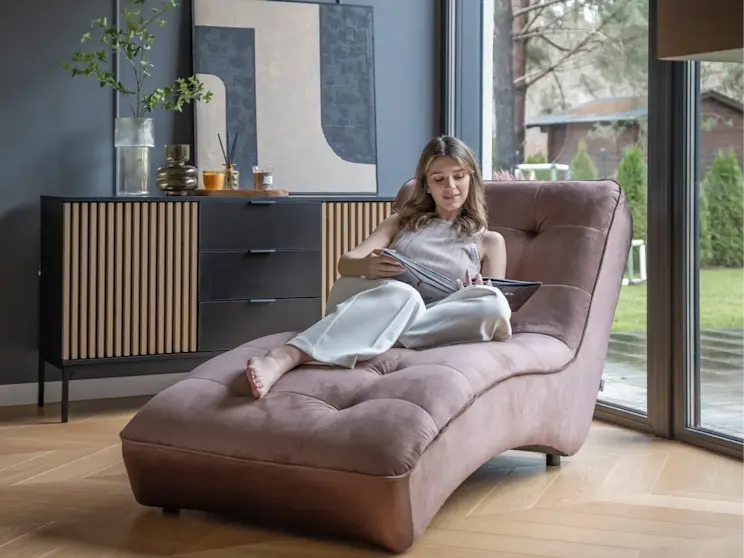 Nowoczesny salon z szezlongiem Loren w ciepłym, pudrowym odcieniu, ustawionym przy dużym przeszkleniu. W tle stoi komoda Meorati z lamelowymi frontami i czarną podstawą, a całość tworzy elegancką strefę relaksu do czytania.