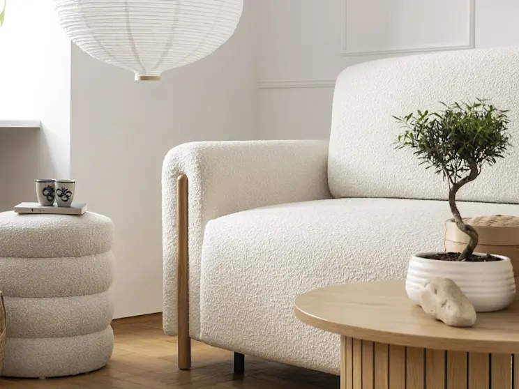Kremowa kanapa Arcadova o miękkiej, strukturalnej tapicerce boucle, ustawiona w jasnym salonie z drewnianą podłogą. W kadrze widać stolik kawowy, puf oraz stonowane dodatki w naturalnych barwach.