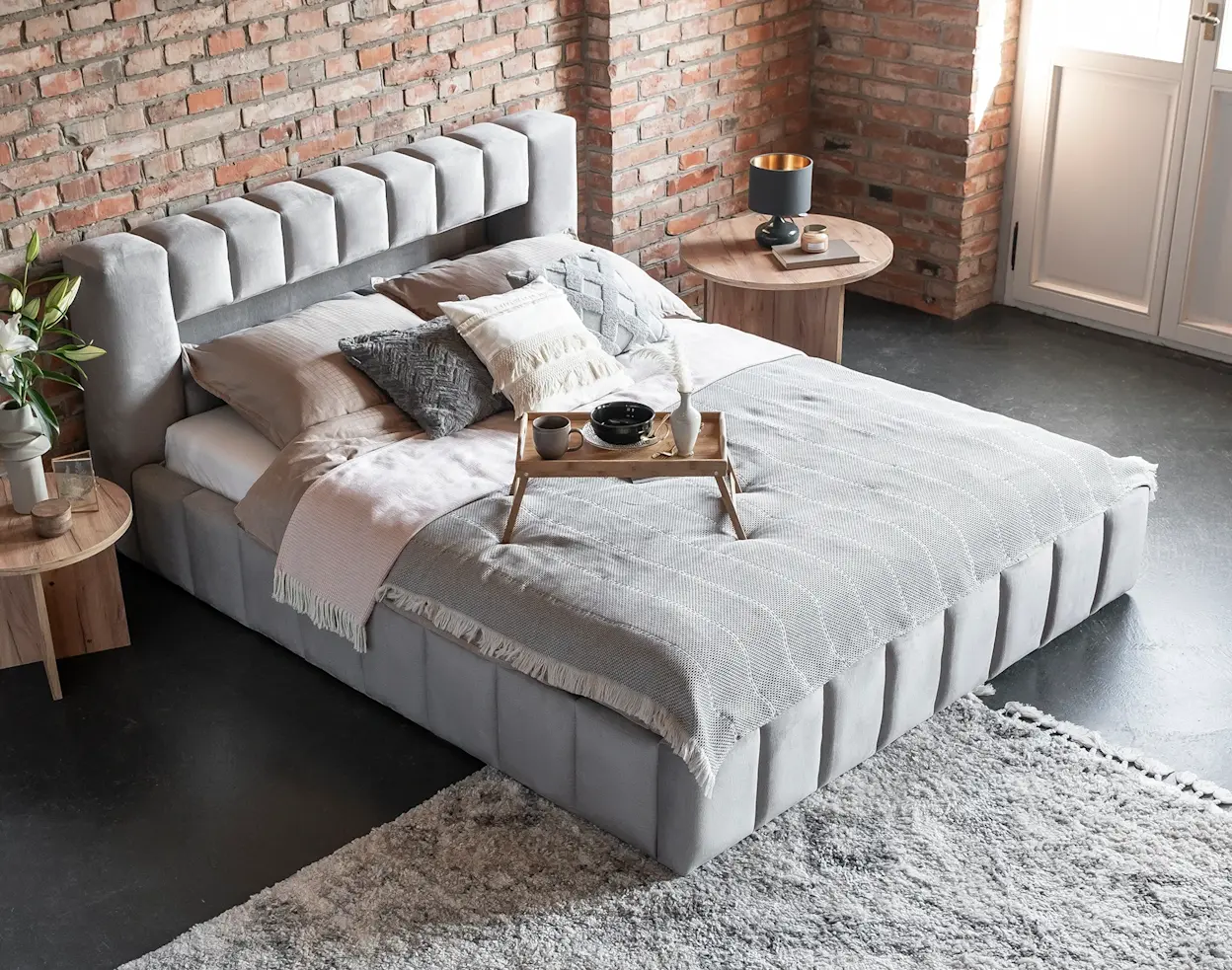 Tapicerowane łóżko Lamica w jasnej sypialni z ceglaną ścianą, z miękkim zagłówkiem i starannie dobranymi tekstyliami.