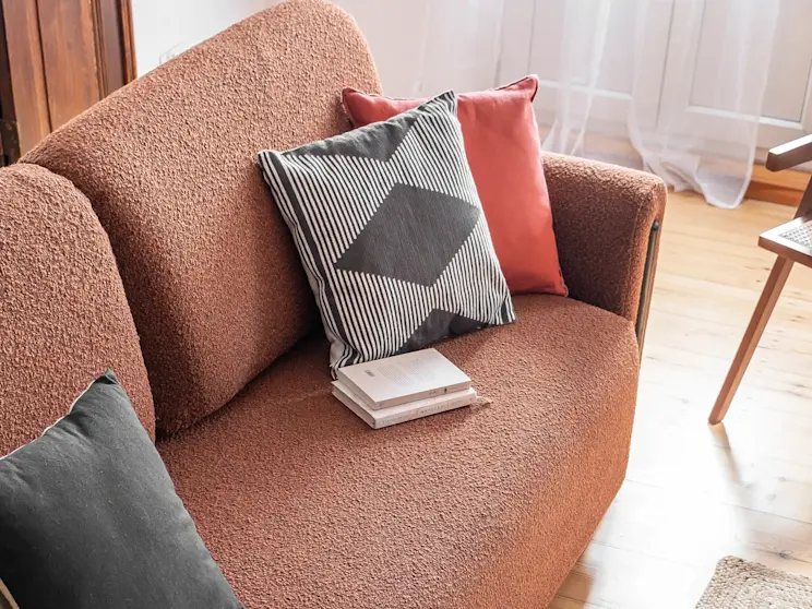 Kanapa Arcadova w tkaninie Jaffray 30, z dekoracyjnymi poduszkami i książkami na siedzisku, w ciepłej, domowej aranżacji.