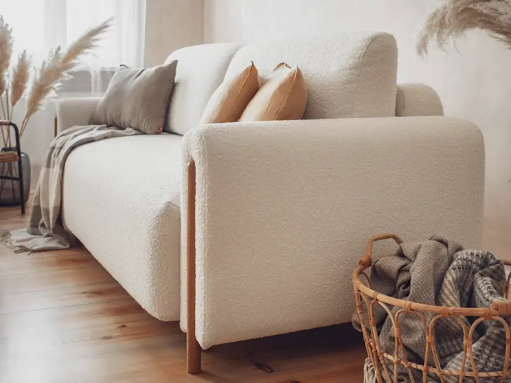 Beżowa kanapa Arcadova z tkaniny boucle, pokazana z boku, ozdobiona poduszkami i pledem, w ciepłej, przytulnej aranżacji z naturalnymi dodatkami.