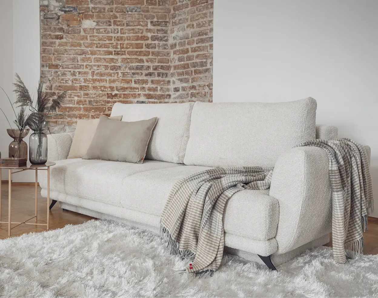 Biała kanapa z funkcją spania Megis obita tkaniną boucle w przytulnej aranżacji salonu. Na siedzisku leżą poduszki i koc w pasy, obok stoi stolik z dekoracjami, a w tle widać ceglaną ścianę i jasny dywan.