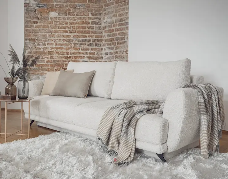 Biała kanapa z funkcją spania Megis obita tkaniną boucle w przytulnej aranżacji salonu. Na siedzisku leżą poduszki i koc w pasy, obok stoi stolik z dekoracjami, a w tle widać ceglaną ścianę i jasny dywan.
