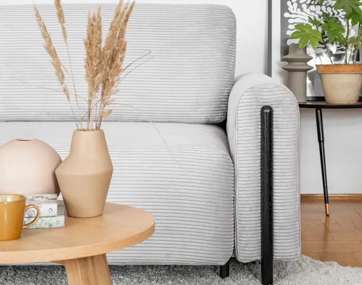 Zbliżenie na kanapę Arcadova II w tkaninie Jarell 03, z dekoracjami w beżowych odcieniach i drewnianym stolikiem w jasnym, przytulnym wnętrzu.