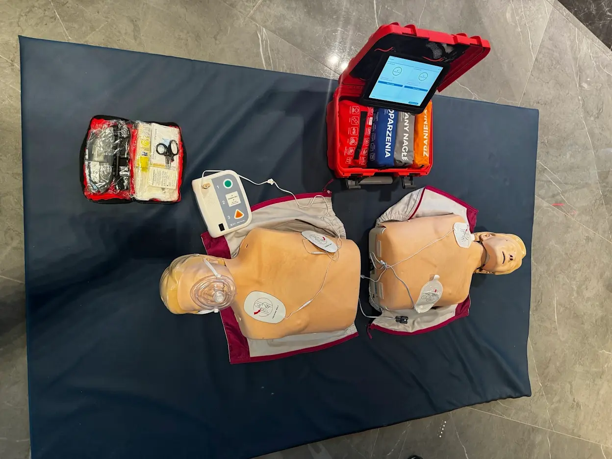 Widok z góry na szkolenie z pierwszej pomocy w ELTAP: dwa fantomy do RKO ułożone na macie z podłączonymi elektrodami. Obok znajduje się defibrylator AED oraz otwarta walizka ratownicza z wyposażeniem i apteczką do ćwiczeń.