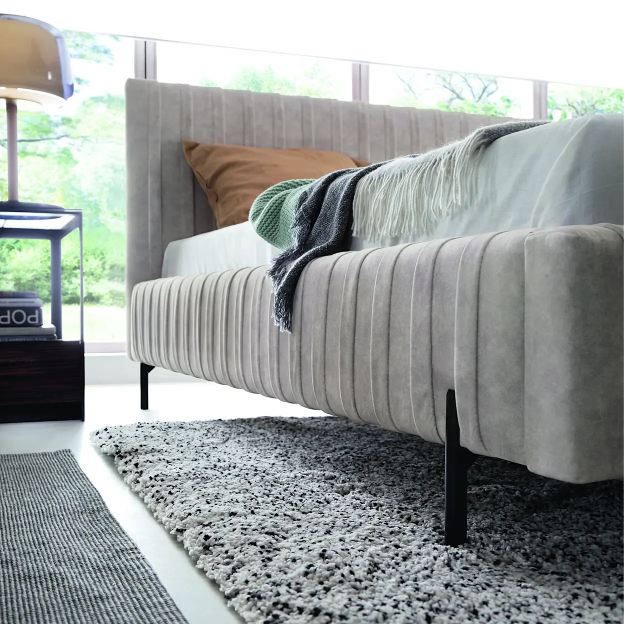 Tapicerowane łóżko Plissa w nowoczesnej sypialni. Zbliżenie na miękką tkaninę, pionowe przeszycia i smukłe czarne nóżki mebla.