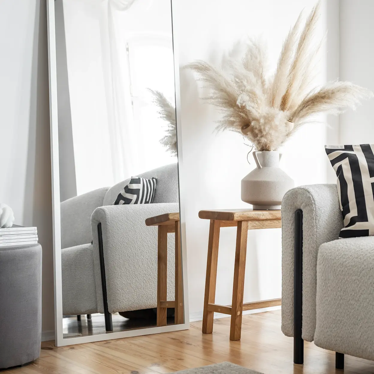 Jasna aranżacja z kanapą Arcadova II, lustrem Medina oraz pufą Linsy, uzupełniona dekoracją z trawy pampasowej i drewnianym stolikiem.