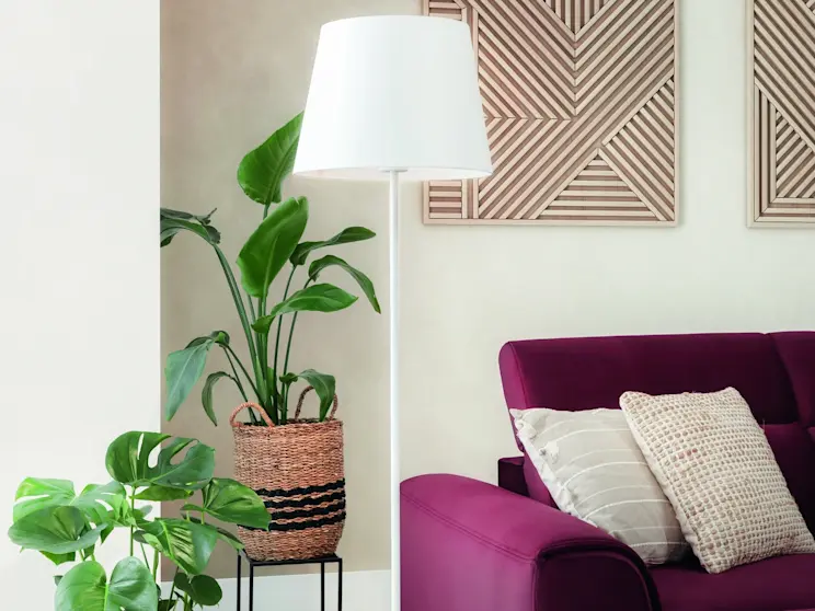 Lampa podłogowa Vespillo w kolorze białym oświetla strefę wypoczynku w salonie. W kadrze widać fioletową sofę z poduszkami, jasną podłogę oraz rośliny doniczkowe w naturalnych, wiklinowych osłonkach.