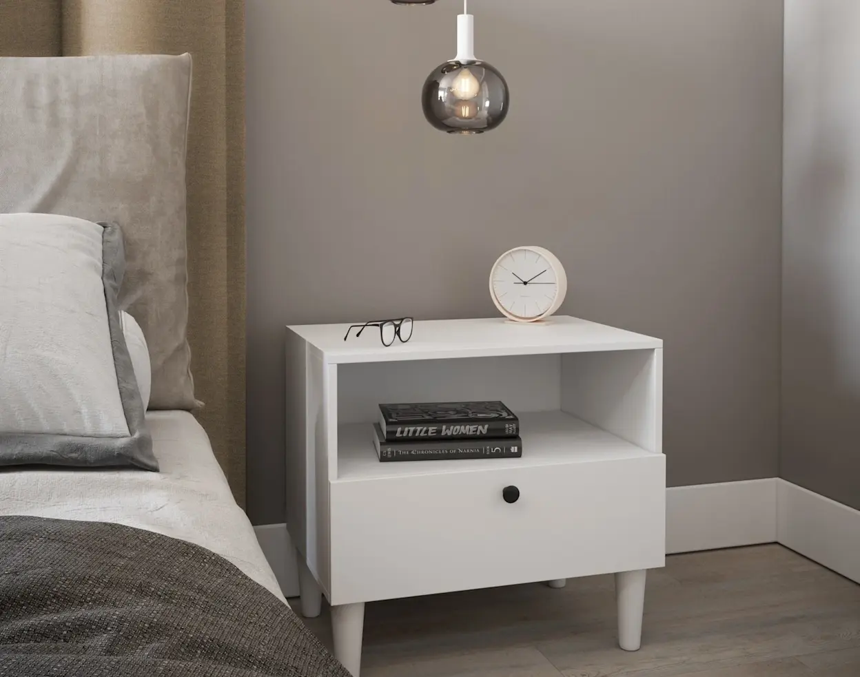 Biały stolik nocny Dullum w nowoczesnej sypialni, ustawiony obok łóżka z tapicerowanym zagłówkiem. Szafka ma szufladę z czarną gałką i otwartą półkę na książki, a na blacie stoją okulary i minimalistyczny zegar.