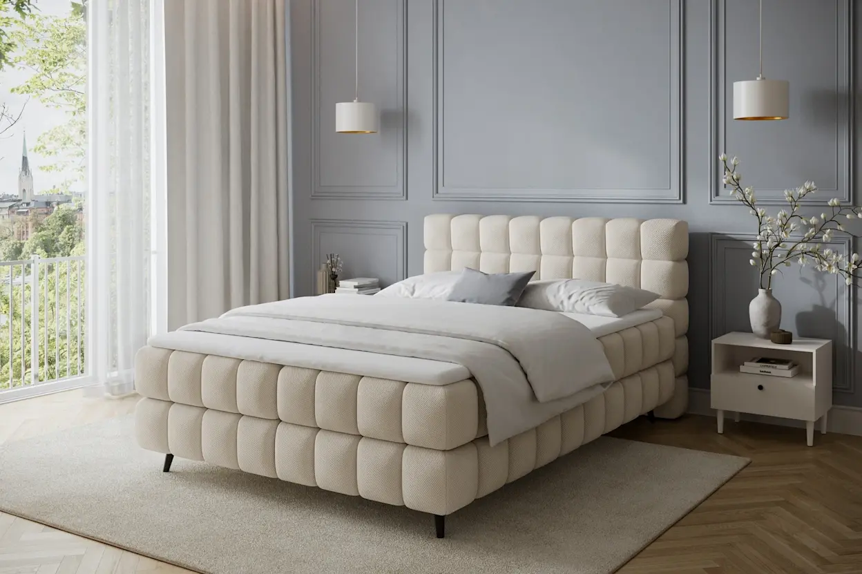 Beżowe łóżko Sabeno Calmora 18 ustawione w eleganckim wnętrzu z drewnianą podłogą, jasnymi zasłonami i stonowaną kolorystyką.