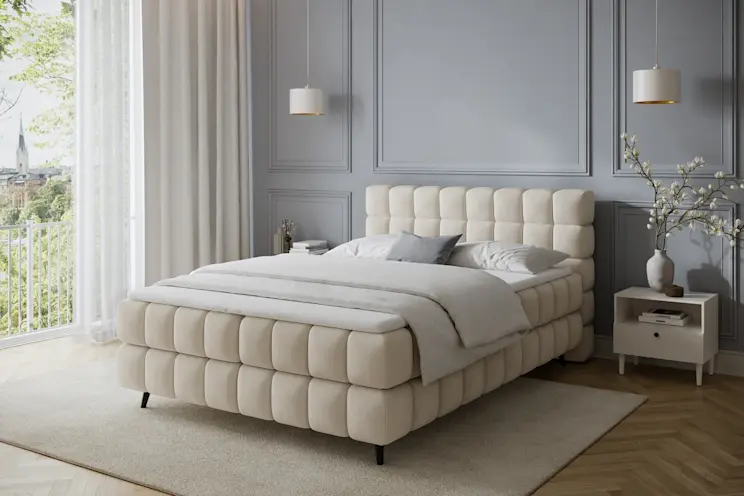 Beżowe łóżko Sabeno Calmora 18 ustawione w eleganckim wnętrzu z drewnianą podłogą, jasnymi zasłonami i stonowaną kolorystyką.