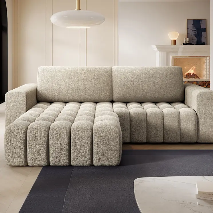 Narożnik Bonett Royal 20 w jasnym beżowym odcieniu, tapicerowany miękką tkaniną typu bouclé. Szezlong z przodu ma wyraźne, pionowe przeszycia, a całość tworzy przytulną strefę wypoczynku w salonie.