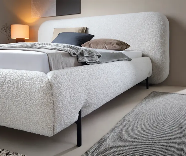 Nowoczesne łóżko Pointt Jaffray 18 z miękką, jasną tapicerką i obłym zagłówkiem obejmującym całą szerokość. Minimalistyczna sypialnia z neutralną pościelą, kocem i poduszkami podkreśla przytulny, elegancki klimat.