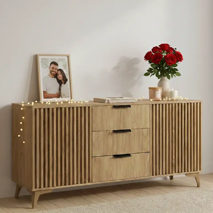 Komoda Entsian w kolorze dąb artisan z ryflowanymi frontami i trzema szufladami. Na blacie stoi wazon z czerwonymi różami, świeca oraz ramka ze zdjęciem, a całość tworzy ciepłą, przytulną aranżację salonu.