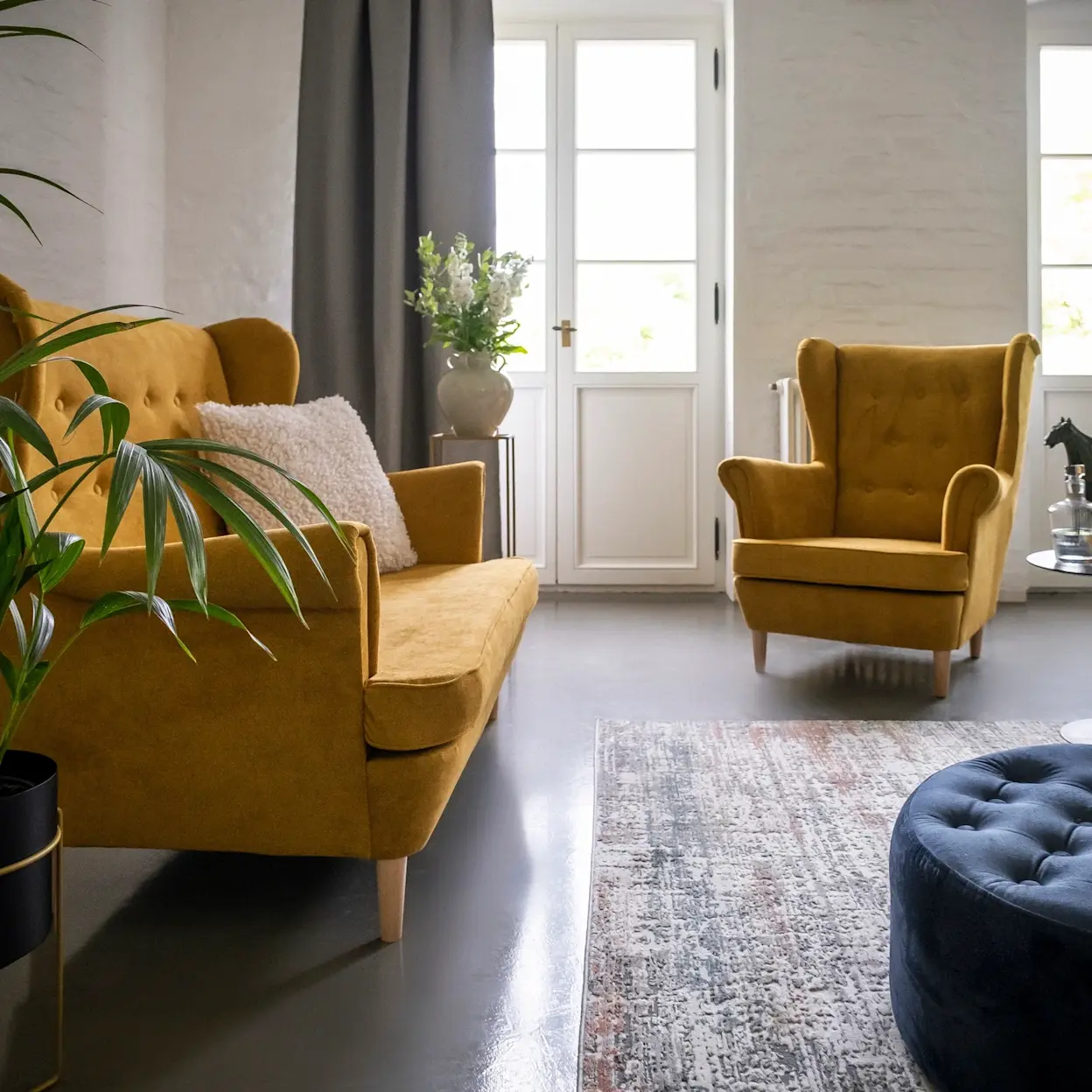 Przytulny salon z musztardową sofą Aros i fotelem wypoczynkowym Aros w tkaninie Omega 68. Na podłodze leży dywan, a z przodu stoi granatowa pufa Monty Monolith 77, całość dopełniają rośliny i jasne okno.