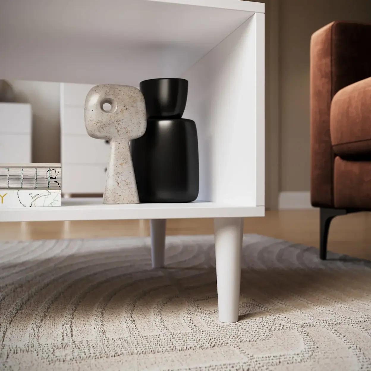 Zbliżenie na dolną półkę stolika kawowego Dullum z dekoracyjnymi figurkami i książką, ustawionego na jasnym, teksturowanym dywanie.