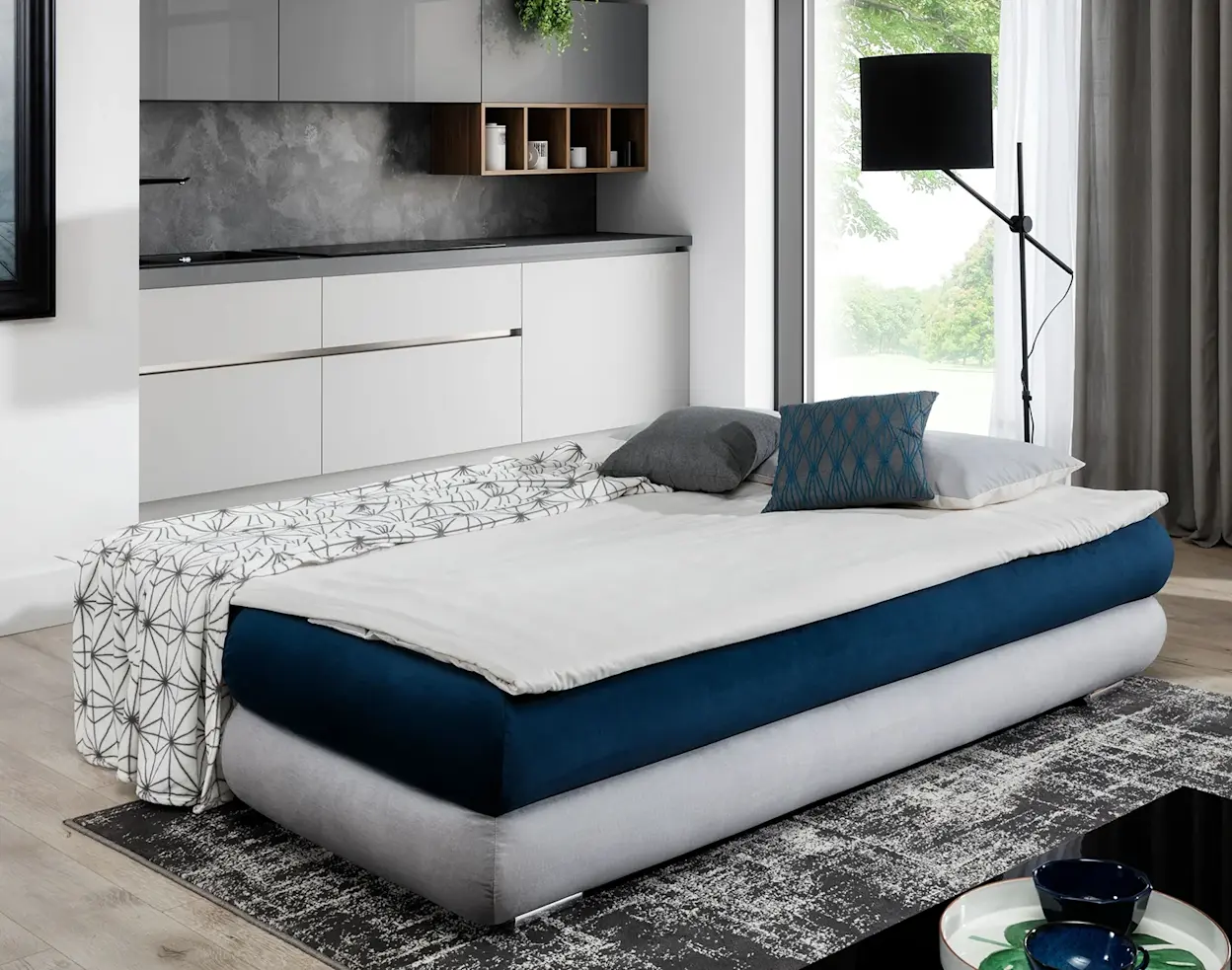 Rozłożona kanapa Milo z funkcją spania w salonie z aneksem kuchennym, z pościelą i poduszkami, przy dużym oknie i zasłonach.