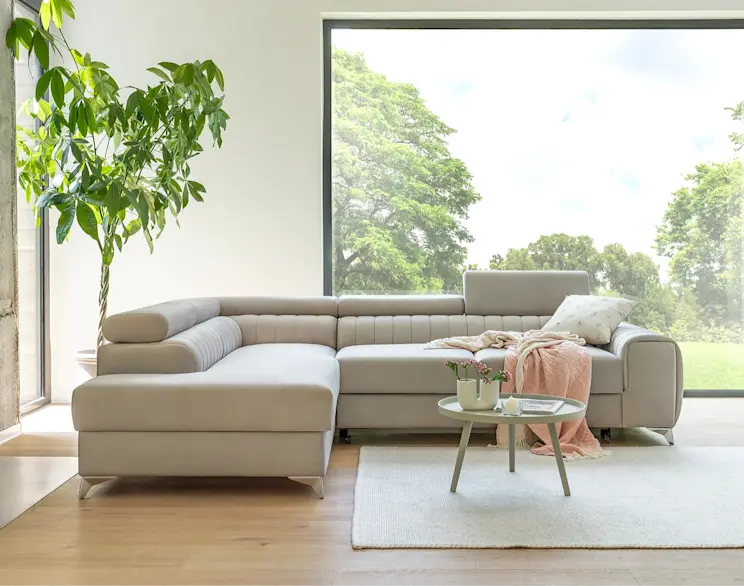 Beżowy narożnik z funkcją spania w jasnym salonie, z podnoszonym zagłówkiem i pikowanym oparciem. Nowoczesny mebel do wypoczynku i codziennego relaksu, idealny do wnętrz w ciepłych kolorach.