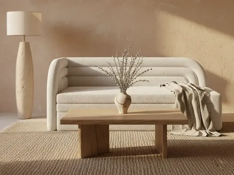 Kremowa kanapa Fabillo ustawiona w jasnym salonie z beżową ścianą o dekoracyjnej fakturze, drewnianym stolikiem kawowym oraz wazonem z gałązkami, tworząca ciepłą i harmonijną aranżację wnętrza w naturalnym stylu.