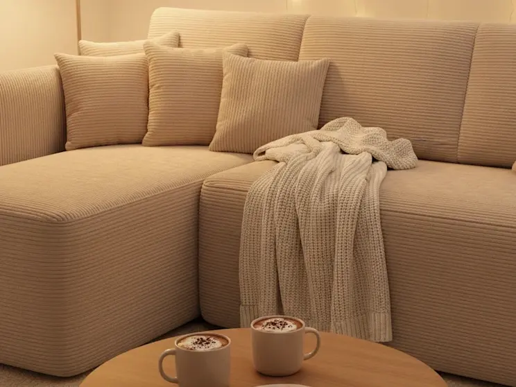 Komfortowy narożnik Hadson w beżowej tkaninie o wyraźnej fakturze. Ciepłe światło lampek i dodatki w neutralnych kolorach tworzą spokojne wnętrze, które sprzyja relaksowi po całym dniu.